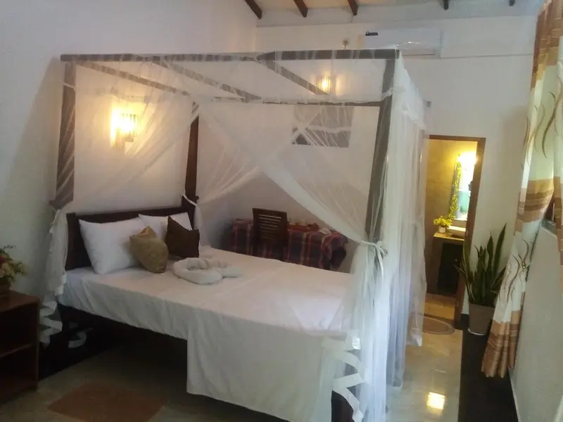 The Double Room with Garden View: a teak four-poster bed with a white mosquito net, wall light and sofa